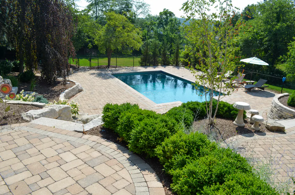 Elevated view of pool with paver deck, planting beds, low stone walls, black fence, lounge chairs, and garden edges